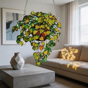 May include: A hanging stained glass plant featuring numerous heart-shaped leaves in shades of yellow, lime green, and deep emerald green. The leaves are connected by dark metal soldering, and the entire piece is suspended by silver-toned chains.