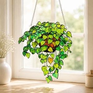 May include: A stained glass hanging plant featuring numerous heart-shaped leaves in various shades of vibrant green and yellow. The leaves are connected by dark metal lines, forming a trailing vine structure suspended by a silver-toned metal chain.