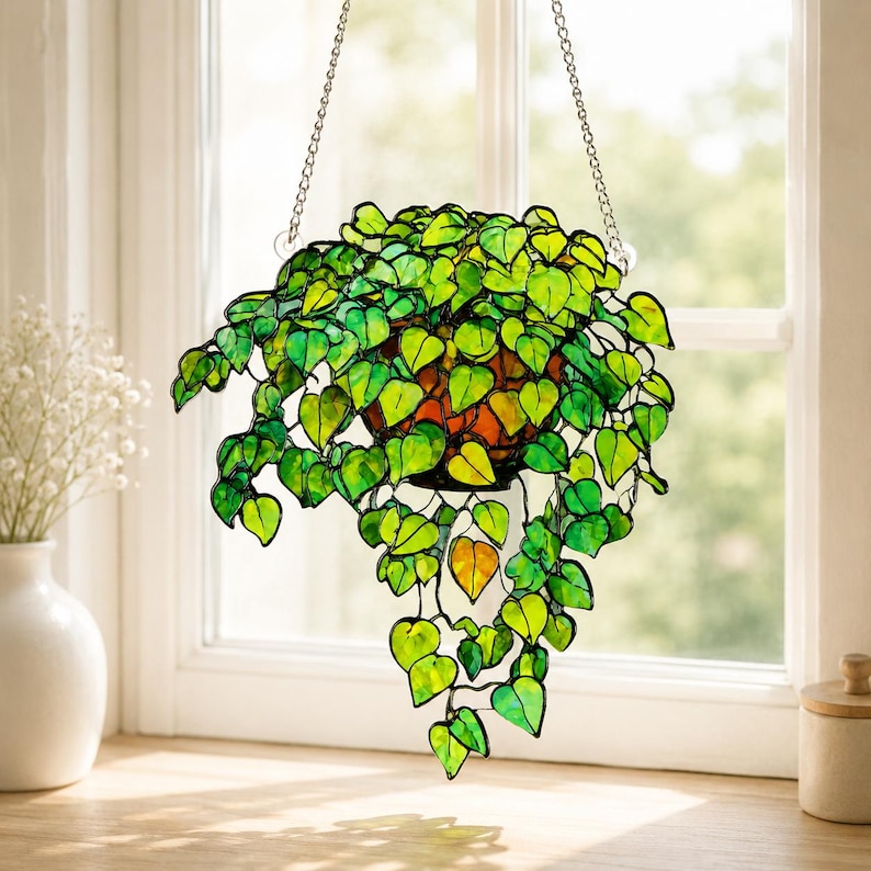 May include: A stained glass hanging plant featuring numerous heart-shaped leaves in various shades of vibrant green and yellow. The leaves are connected by dark metal lines, forming a trailing vine structure suspended by a silver-toned metal chain.