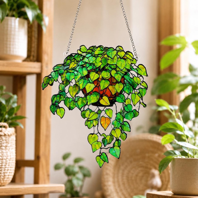 May include: A hanging stained glass suncatcher shaped like a potted trailing plant with vibrant green heart-shaped leaves and a single orange leaf. The piece is constructed with dark metal soldering and is suspended by two silver-toned metal chains.