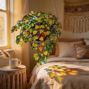 May include: A hanging stained glass plant sculpture featuring numerous heart-shaped leaves in shades of green and yellow. The piece is suspended by silver-toned metal chains and casts colourful light patterns onto the bedding below it.
