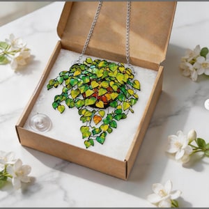 May include: A stained glass suncatcher featuring a potted plant with vibrant green and yellow heart-shaped leaves, suspended by a silver metal chain. The piece is packaged in a brown cardboard box with a clear suction cup included for display.