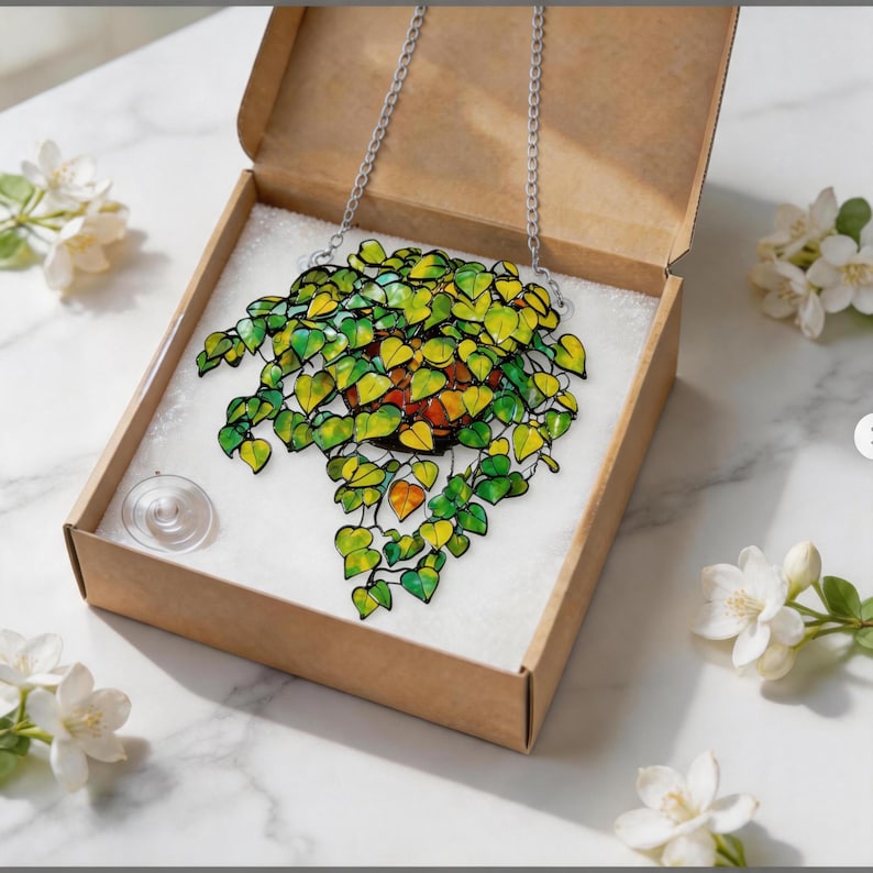 May include: A stained glass suncatcher featuring a potted plant with vibrant green and yellow heart-shaped leaves, suspended by a silver metal chain. The piece is packaged in a brown cardboard box with a clear suction cup included for display.