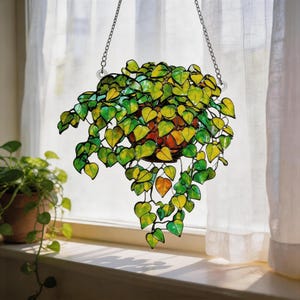May include: A hanging stained glass plant sculpture featuring numerous heart-shaped leaves in shades of vibrant green and yellow. The leaves are connected by dark metal solder lines, forming a cascading vine effect suspended by two silver metal chains.