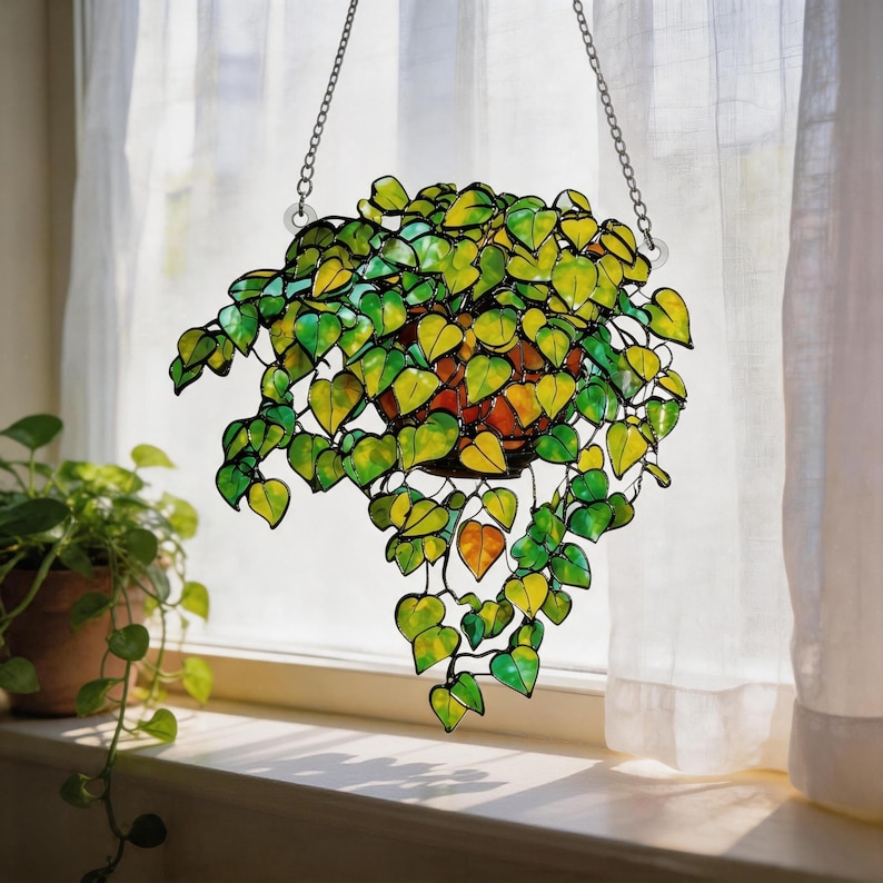 May include: A hanging stained glass plant sculpture featuring numerous heart-shaped leaves in shades of vibrant green and yellow. The leaves are connected by dark metal solder lines, forming a cascading vine effect suspended by two silver metal chains.