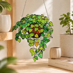 May include: A stained glass hanging plant ornament featuring numerous heart-shaped leaves in various shades of vibrant green and yellow. The leaves are connected by thin black metal lines, and the piece is suspended by a silver-toned metal chain.