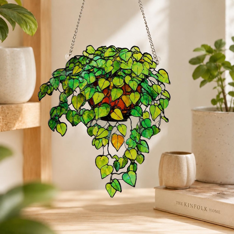 May include: A stained glass hanging plant ornament featuring numerous heart-shaped leaves in various shades of vibrant green and yellow. The leaves are connected by thin black metal lines, and the piece is suspended by a silver-toned metal chain.