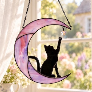 Pode incluir: Um apanhador de sol de vitral em forma de lua crescente em tons de rosa e roxo, apresentando a silhueta de um gato preto sentado na lua e alcançando uma pequena estrela iridescente pendurada por uma corrente de metal preta.