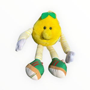 May include: A plush toy shaped like a yellow pear with a smiling face, green leaf accents, and striped arms and legs. The toy has green and white sneakers with orange and red details. The toy is designed to look like a cartoon character.