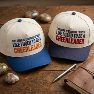 May include: Two off-white baseball caps with blue brims and a dark blue button on top. The caps feature embroidered text that reads: This drink is starting to taste like I used to be a cheerleader, with the word cheerleader in orange or red.