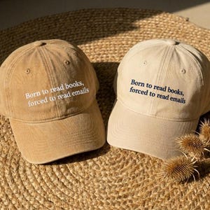 May include: Two baseball caps in tan and light beige, each featuring the embroidered text Born to read books, forced to read emails. The tan cap has white lettering, while the light beige cap features dark navy blue embroidered text.