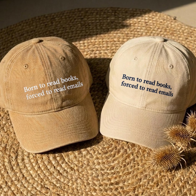 May include: Two baseball caps in tan and light beige, each featuring the embroidered text Born to read books, forced to read emails. The tan cap has white lettering, while the light beige cap features dark navy blue embroidered text.