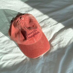May include: A coral-colored baseball cap with a curved brim and a button on top. The text "Favorite Daughter" is embroidered in a dark red script font across the front panels of the cap, which features a six-panel construction with eyelets.