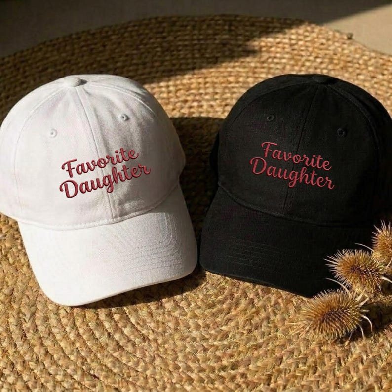 May include: A white baseball cap and a black baseball cap, both featuring the text "Favorite Daughter" embroidered in red script lettering on the front panels. Each cap has a structured crown, a curved brim, and a button on the top center.