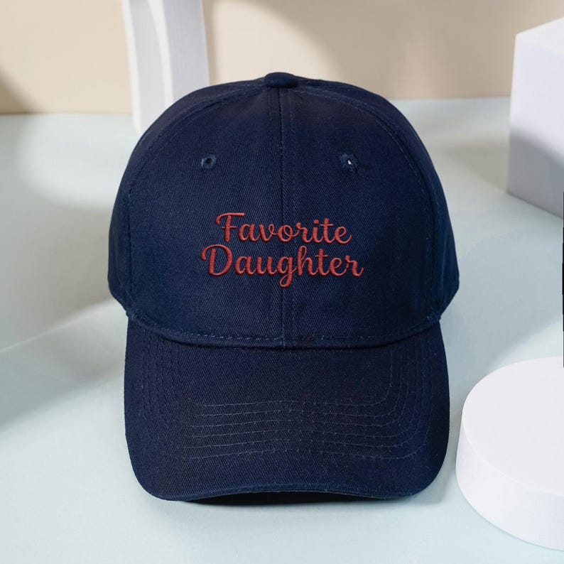 May include: A navy blue baseball cap featuring the text Favorite Daughter embroidered in red thread on the front panel. The cap has a structured crown, a curved brim with visible stitching, and eyelets for ventilation on the upper panels.