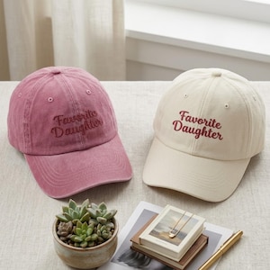 May include: Two baseball caps, one in a dusty rose pink and one in an off-white cream color. Both caps feature the embroidered text "Favorite Daughter" in a dark red script font. Each hat has a structured crown, a curved brim, and eyelets.