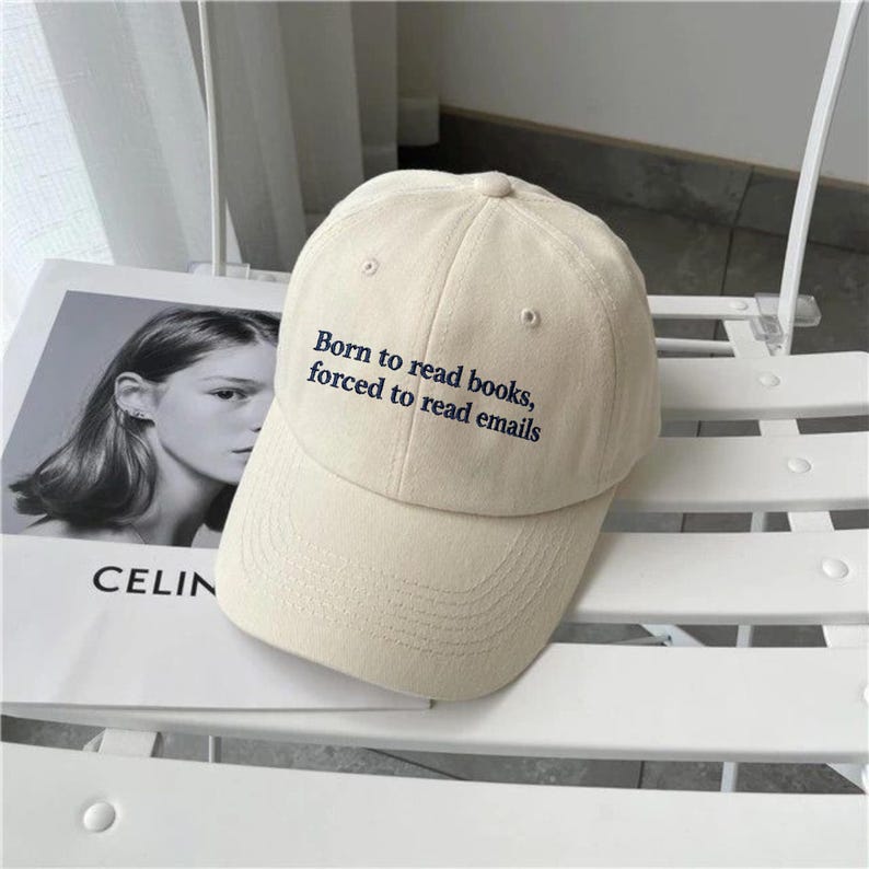 May include: A cream-colored baseball cap featuring the embroidered black text Born to read books, forced to read emails on the front panel. The cap has a structured crown, a curved brim, and a button at the top center of the fabric dome.