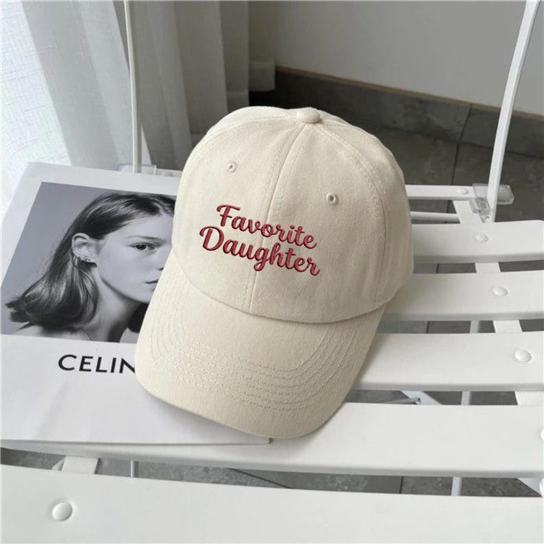 May include: A cream-colored baseball cap featuring the embroidered text Favorite Daughter in a dark red script font. The cap has a structured crown, a curved brim, and small ventilation eyelets on the top panels of the fabric construction.