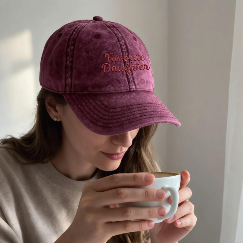 May include: A maroon baseball cap with a washed, vintage-style finish. The front of the cap features the embroidered text "Favorite Daughter" in a cursive, reddish-orange font. The cap has a structured crown, a curved brim, and a button on top.