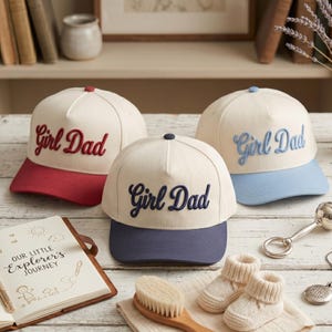 May include: Three baseball caps with "Girl Dad" embroidered on the front. The caps have different color schemes: red bill and lettering, blue bill and lettering, and navy bill with navy lettering. A baby book, booties, and a brush are also in the image.