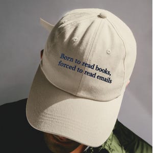 May include: A beige baseball cap featuring the embroidered text Born to read books, forced to read emails in a dark blue serif font. The cap has a structured crown, a curved brim, and a button on top, with a fabric strap visible at the back.