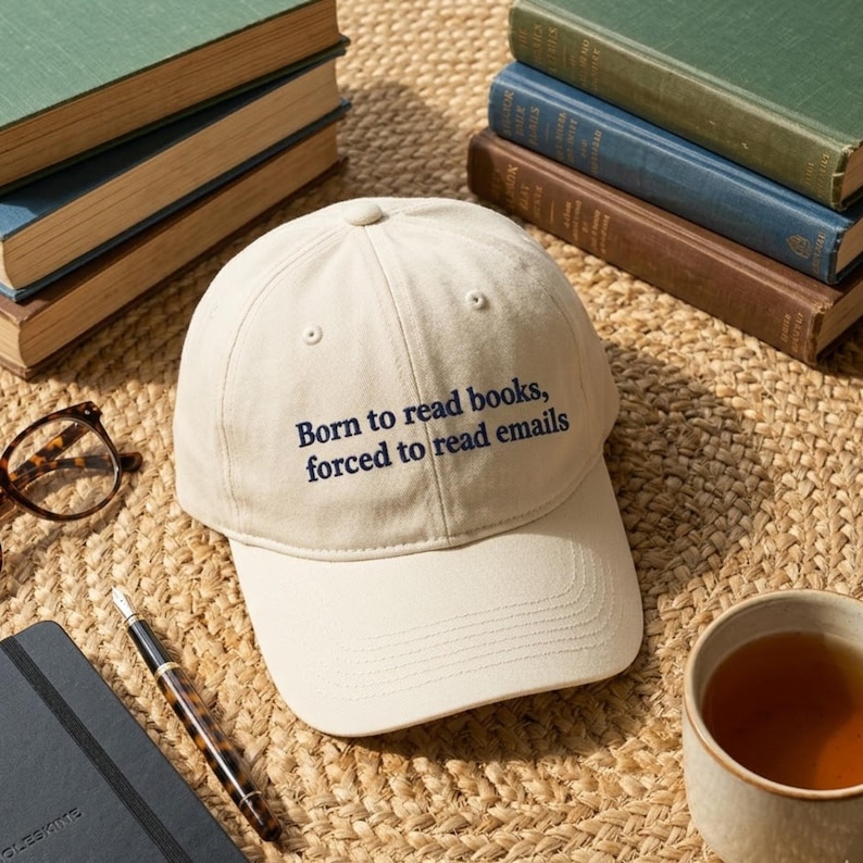 May include: A cream-colored baseball cap featuring the embroidered dark blue text "Born to read books, forced to read emails" on the front panel. The cap has a structured crown, a curved brim, and small ventilation eyelets on the top panels.