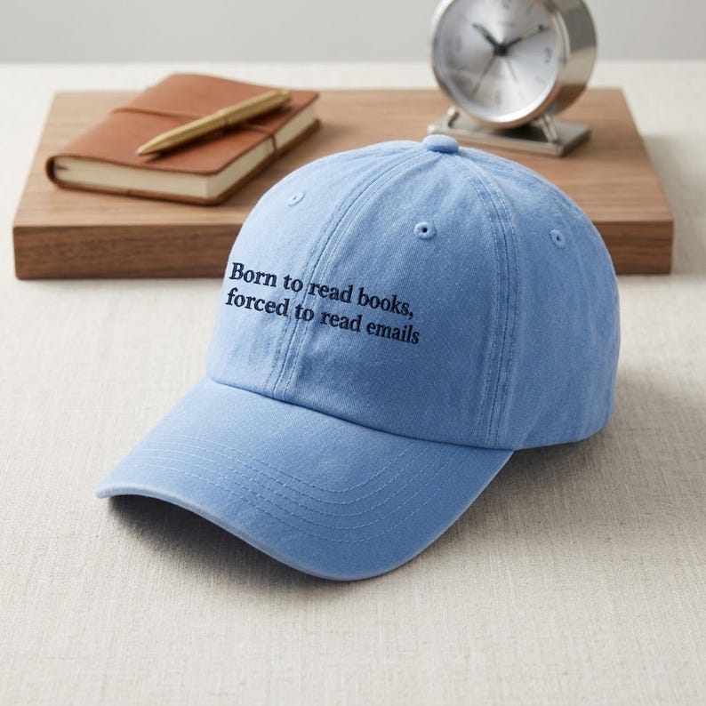 May include: A light blue baseball cap featuring the embroidered black text Born to read books, forced to read emails. The cap has a structured crown, a curved brim, and stitched eyelets on the top panels for ventilation and a classic design.