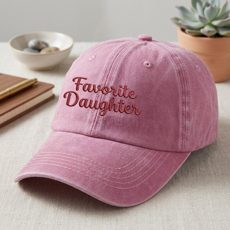 May include: A dusty rose baseball cap with a six-panel construction and a curved brim. The front of the cap features the embroidered text "Favorite Daughter" in a dark red, cursive script font. The fabric has a soft, washed-out texture.
