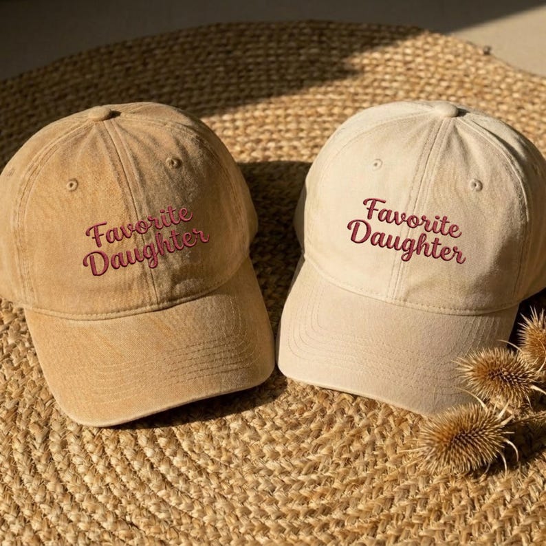 May include: Two baseball caps in tan and beige shades, each featuring the embroidered text "Favorite Daughter" in a dark red script font. The caps have a structured crown, a curved brim, and eyelets on the top panels for ventilation.