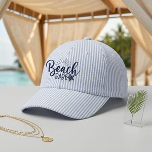 May include: A light blue and white striped baseball cap with the text "Beach Babe" in navy blue script. The cap has a curved brim and a seersucker texture, perfect for beach outings. A gold necklace is in the foreground.