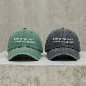 May include: Two baseball caps, one in a washed green and one in a washed charcoal gray, both featuring the white embroidered text Born to read books, forced to read emails on the front panel. Each cap has a curved brim and a six-panel construction.