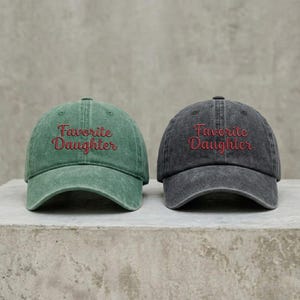 May include: Two baseball caps with a distressed finish, one in sage green and one in charcoal gray. Each cap features the text "Favorite Daughter" embroidered in red cursive script across the front panel, with a curved brim and a structured crown.
