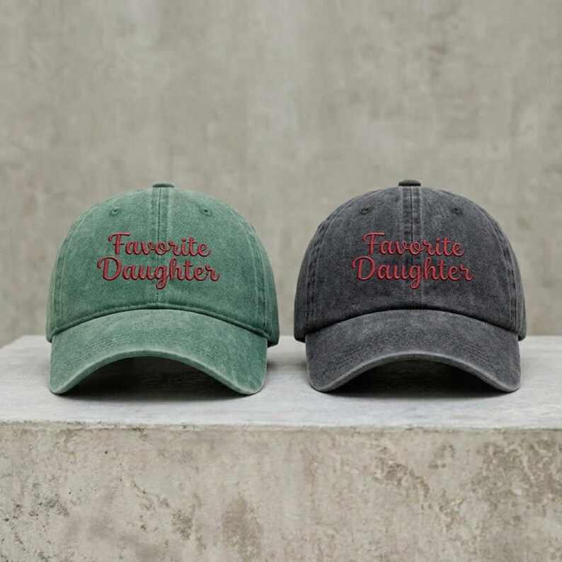 May include: Two baseball caps with a distressed finish, one in sage green and one in charcoal gray. Each cap features the text "Favorite Daughter" embroidered in red cursive script across the front panel, with a curved brim and a structured crown.