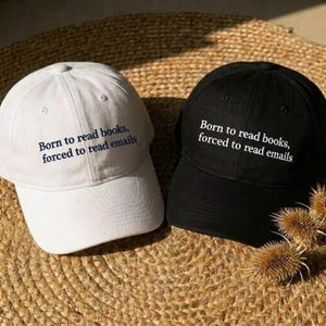 May include: A white baseball cap with dark blue text and a black baseball cap with white text. Both caps feature the embroidered phrase: Born to read books, forced to read emails. Each cap has a structured crown, a curved brim, and eyelets.