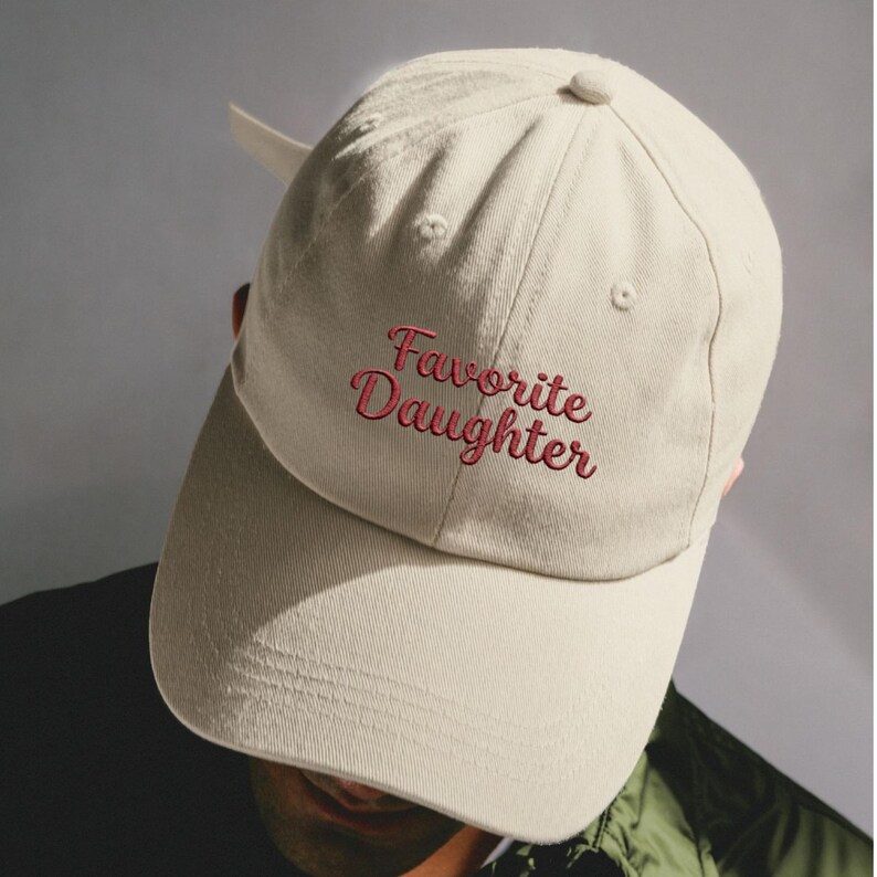 May include: A beige baseball cap featuring the embroidered text Favorite Daughter in a dark red cursive font. The cap has a structured crown, a curved brim, and a button on top, with a fabric strap visible at the back for an adjustable fit.