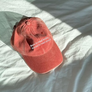 May include: A coral-colored baseball cap with a six-panel construction and a curved brim. The front of the cap features white embroidered text that reads, Born to read books, forced to read emails, centered above the brim on the crown.