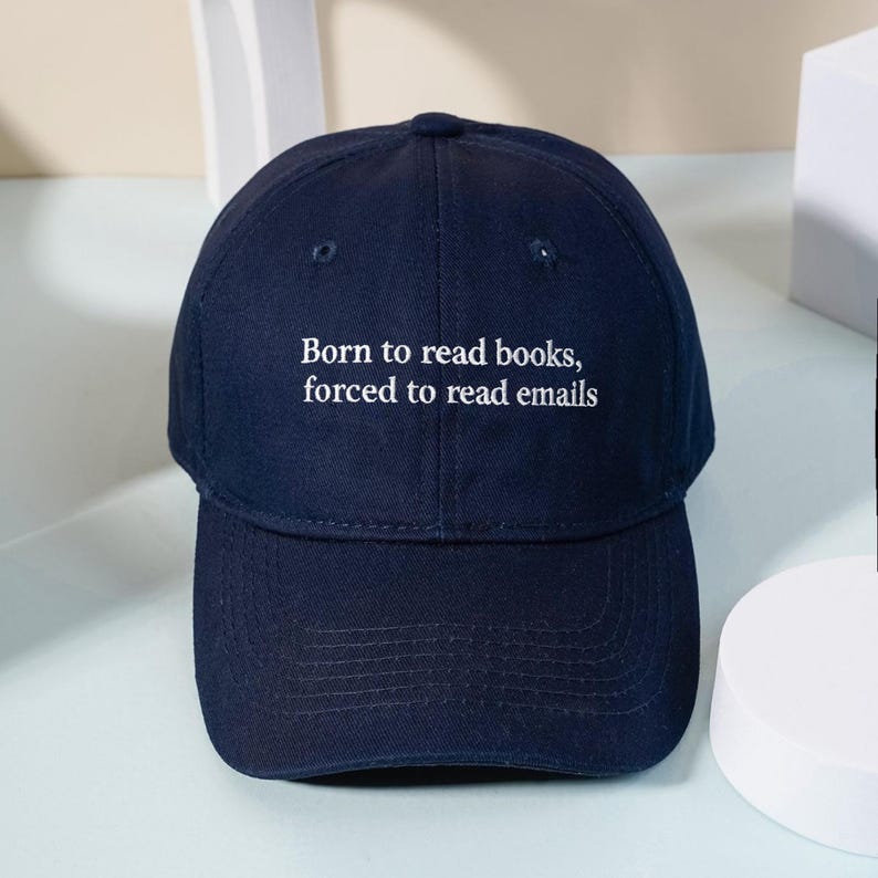 May include: A navy blue baseball cap featuring the white embroidered text Born to read books, forced to read emails centered on the front panel. The cap has a structured crown, a curved brim, and stitched eyelets for ventilation on the top panels.