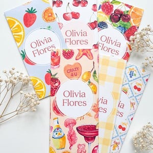 Può includere: Una collezione di segnalibri colorati con illustrazioni di frutta e bevande. Ogni segnalibro presenta il nome "Olivia Flores" in un design circolare. I disegni includono fragole, ciliegie e cocktail.