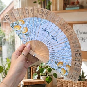 Puede incluir: Abanico de mano de madera con un dise&ntilde;o estampado en azul claro y blanco con limones. El abanico tiene un dise&ntilde;o calado decorativo a lo largo del borde superior y el texto "Julia y Daniel".