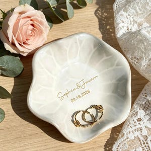 Puede incluir: Un plato de cerámica blanco con diseño de hojas, que contiene dos anillos de oro. El plato está inscrito con los nombres "Sophia & Jacson" y la fecha "09.16.2026". Una rosa rosa y vegetación en el fondo.