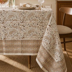 May include: A beige tablecloth with a brown paisley pattern and a decorative border. The tablecloth is draped over a wooden table, with a portion of the cloth hanging off the edge. The table is set with plates, napkins, and glassware.