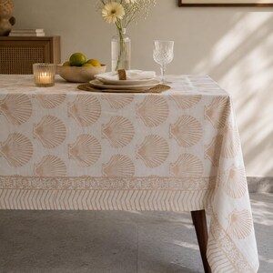 Puede incluir: Un mantel beige con un estampado de conchas cubre una mesa de madera. La mesa está puesta con platos, vasos y un jarrón con flores. Una vela y un cuenco de fruta también están sobre la mesa. El mantel tiene un borde decorativo.