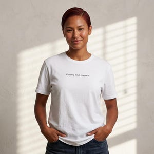 May include: A white t-shirt with the phrase "Raising kind humans" printed in black. The shirt is short-sleeved and appears to be made of a soft material. The person wearing the shirt is smiling.