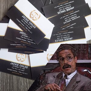 May include: A stack of black and white business cards with gold accents. The business cards feature the name "Jackie Chiles" and the text "Attorney at Law". The text "Your face is my case!" is also on the cards. The business cards are stacked on a wooden surface.