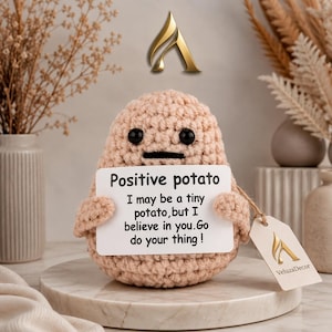Peut inclure: Une figurine de pomme de terre crochetée beige avec des yeux en perles noires et une bouche noire plate. Elle tient un panneau rectangulaire blanc avec le texte : Positive potato. I may be a tiny potato, but I believe in you. Go do your thing!