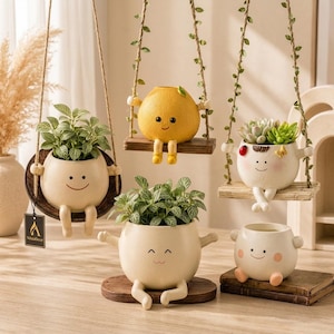 May include: A collection of round ceramic planters with smiling faces and dangling legs. Three planters hang from ropes on wooden swings, while two sit on surfaces. The planters feature green plants, including a succulent and leafy foliage.