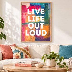 Op de afbeelding: Een rechthoekige wandprint in een lichthouten lijst met de witte tekst LIVE LIFE OUT LOUD gecentreerd over een levendige, abstracte achtergrond van gemengde penseelstreken in tinten magenta, oranje, geel, blauw en donkerpaars.