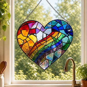 Puede incluir: Un corazón de vidriera en colores del arcoíris, con un marco negro, colgado de una cadena. El corazón presenta un espectro de colores, incluyendo rojo, naranja, amarillo, verde, azul y morado. El arte de la ventana se exhibe frente a una ventana.