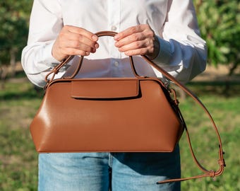 May include: A structured brown leather doctor-style handbag featuring a wide rectangular frame, a top handle, and a thin adjustable shoulder strap. The bag has a smooth finish, a flap closure, and a wide base, designed for carrying personal items.