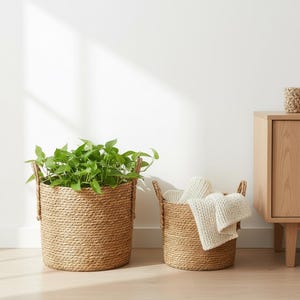 Puede incluir: Dos cestas cilíndricas tejidas. Una cesta contiene una planta verde, la otra tiene mantas blancas dobladas. Ambas cestas tienen asas. Las cestas están hechas de un material marrón claro texturizado.